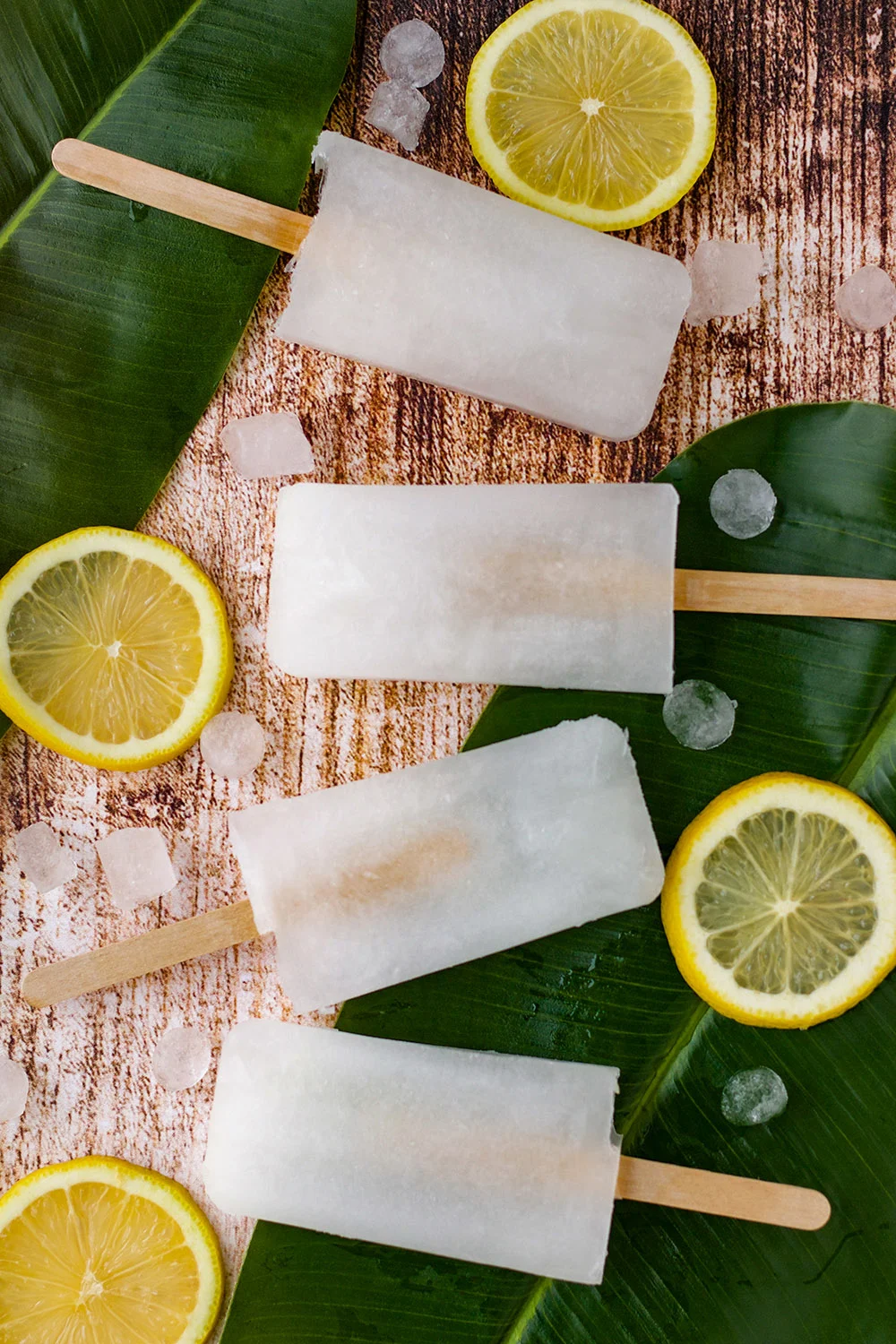 Four ice pops on a table with ice cubes, leaves, and lemon slices.