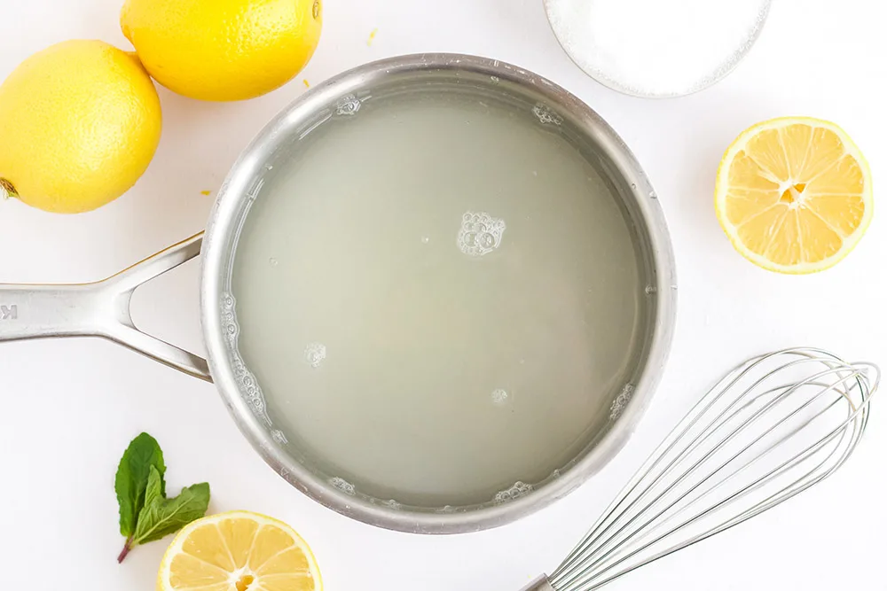 Water and sugar in a pan surrounded by lemons, sugar, and a whisk. 