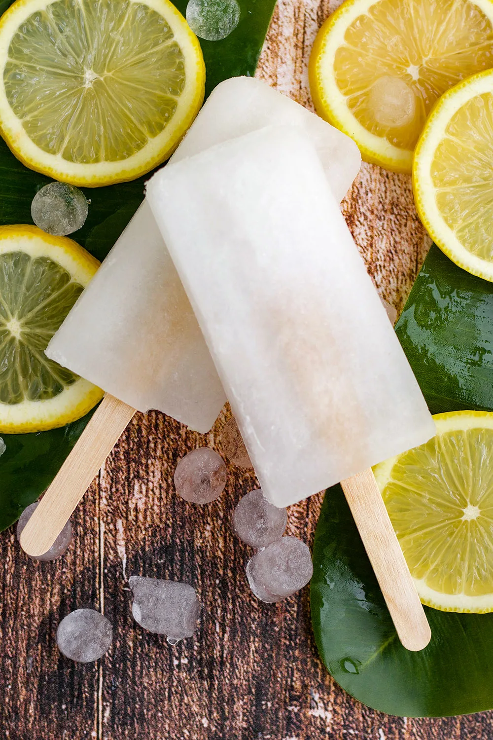 Two lemon pops on top of each other with lemon slices and nugget ice on the table.