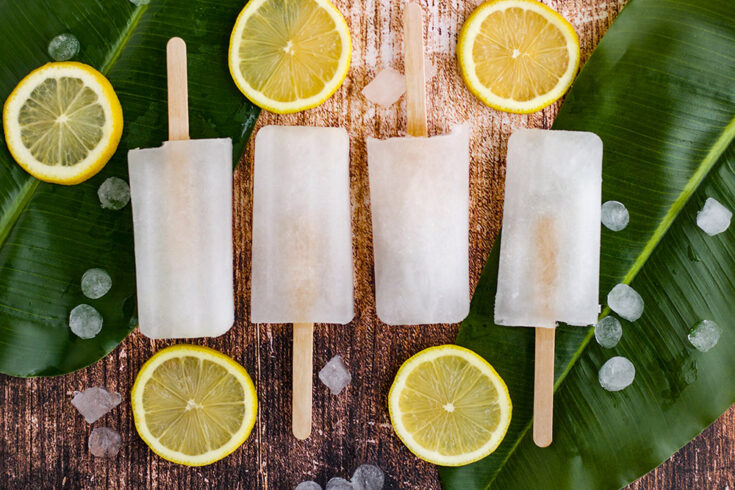 Four lemon popsicles in a row on a table with greens and lemon slices.