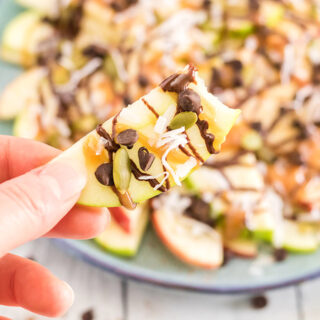 Holding an apple nacho above a plate of apple slices with toppings.