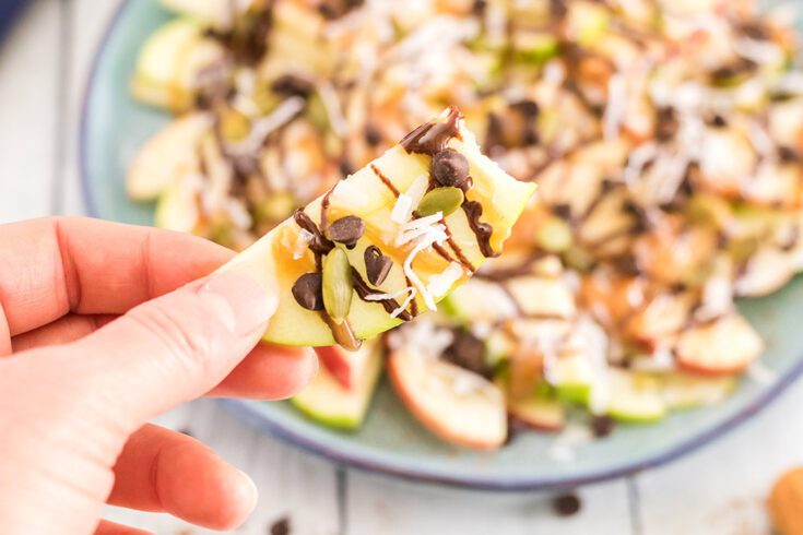 Holding an apple nacho above a plate of apple slices with toppings.