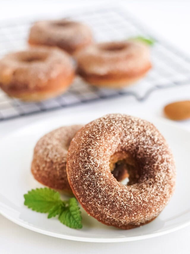 Two apple donuts on a plate with more on a rack in the background.