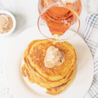 Pouring syrup over pumpkin pancakes with a scoop of cinnamon butter.