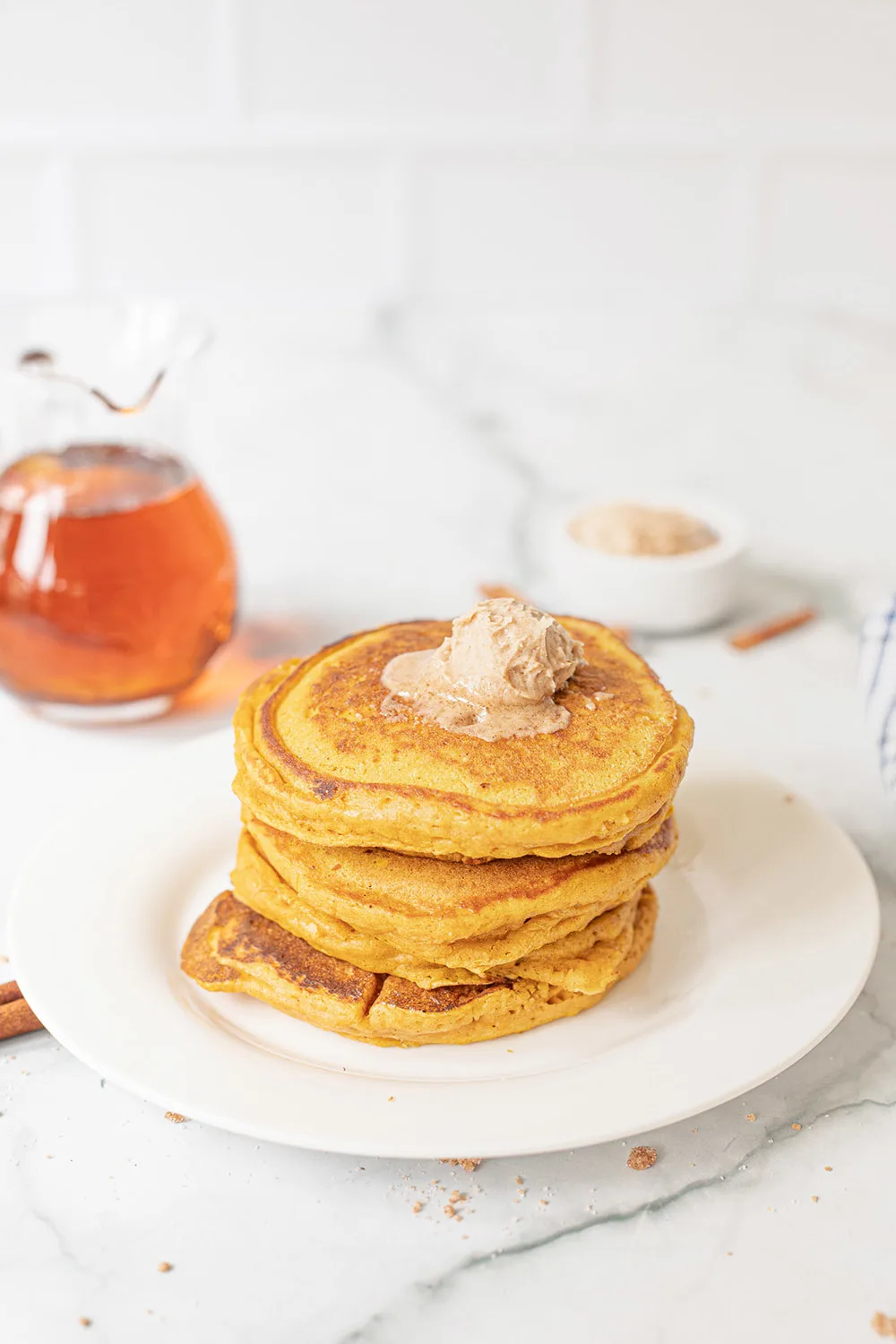 Pancakes on a plate topped with honey butter and a pitcher of syrup in the background.