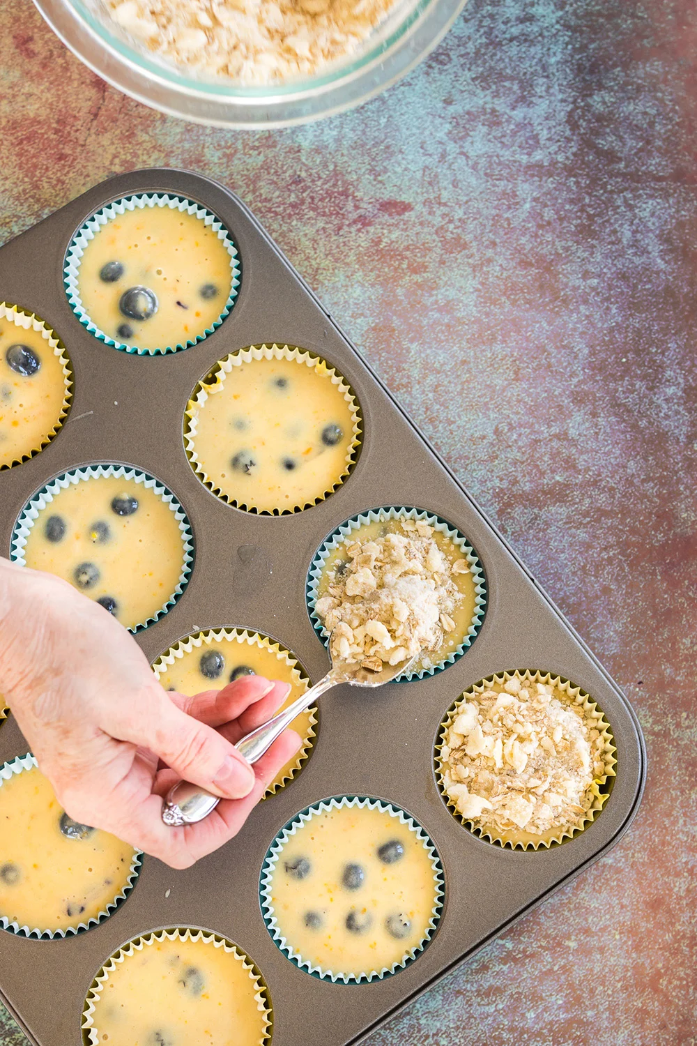 Adding topping to blueberry muffins in a tin.