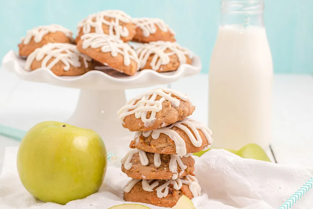 Stacked apple cookies next to a green apple and a plate of more cookies.