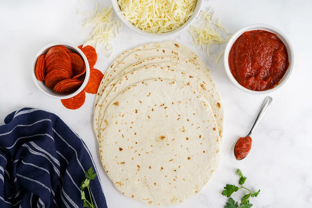 Tortillas, cheese, pepperoni, and sauce on a table.