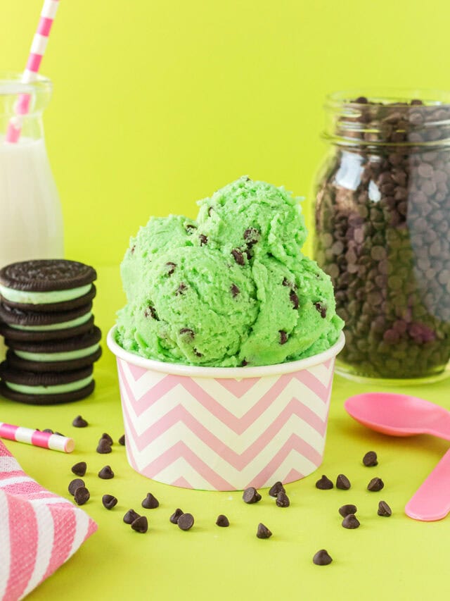 Pink cup of mint chip cookie dough with cookies, milk, pink spoons, and chips on a green background.