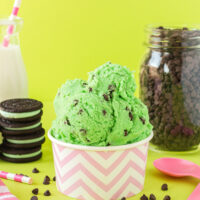 Pink cup of mint chip cookie dough with cookies, milk, pink spoons, and chips on a green background.