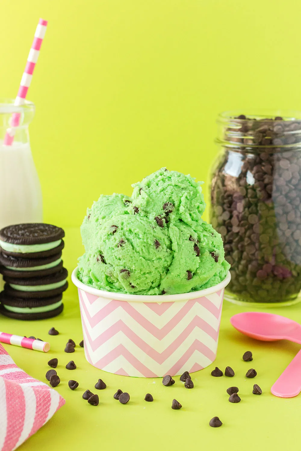 Pink cup of mint chip cookie dough with cookies, milk, pink spoons, and chips on a green background.