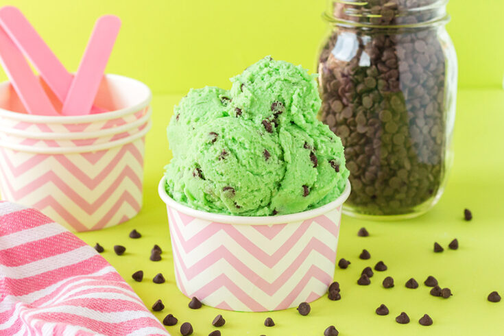 Scoops of mint chip cookie dough in a pink striped cup with chips on the table.
