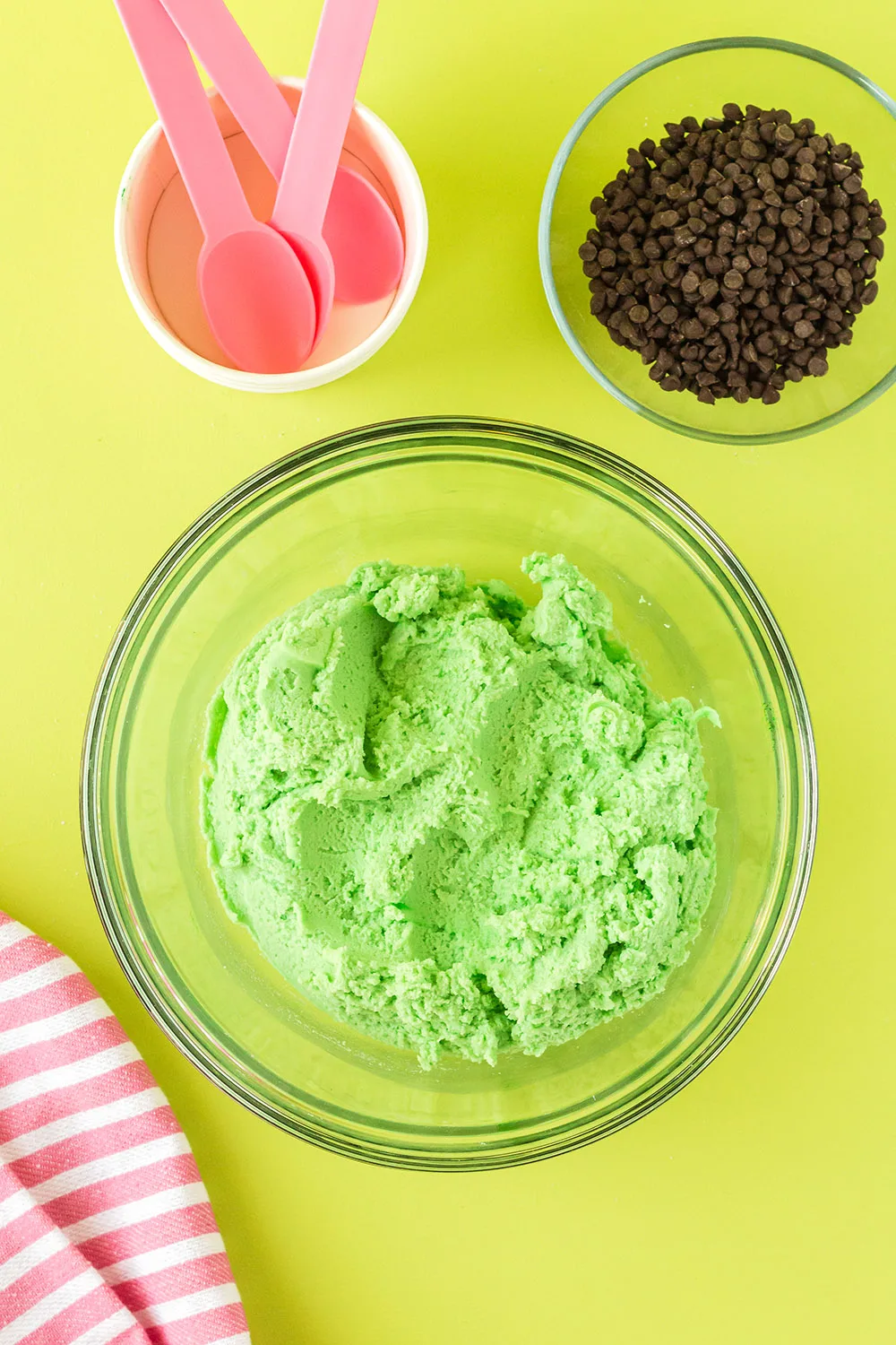 Green cookie dough in a mixing bowl next to chips and pink spoons. 