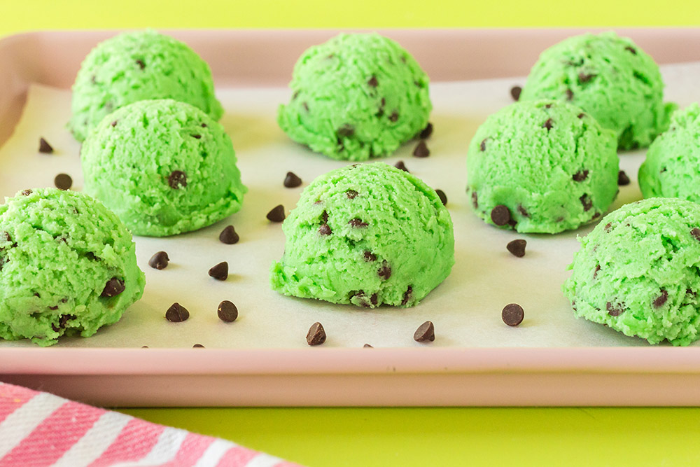 Scoops of green dough with mini chocolate chips on baking sheet. 