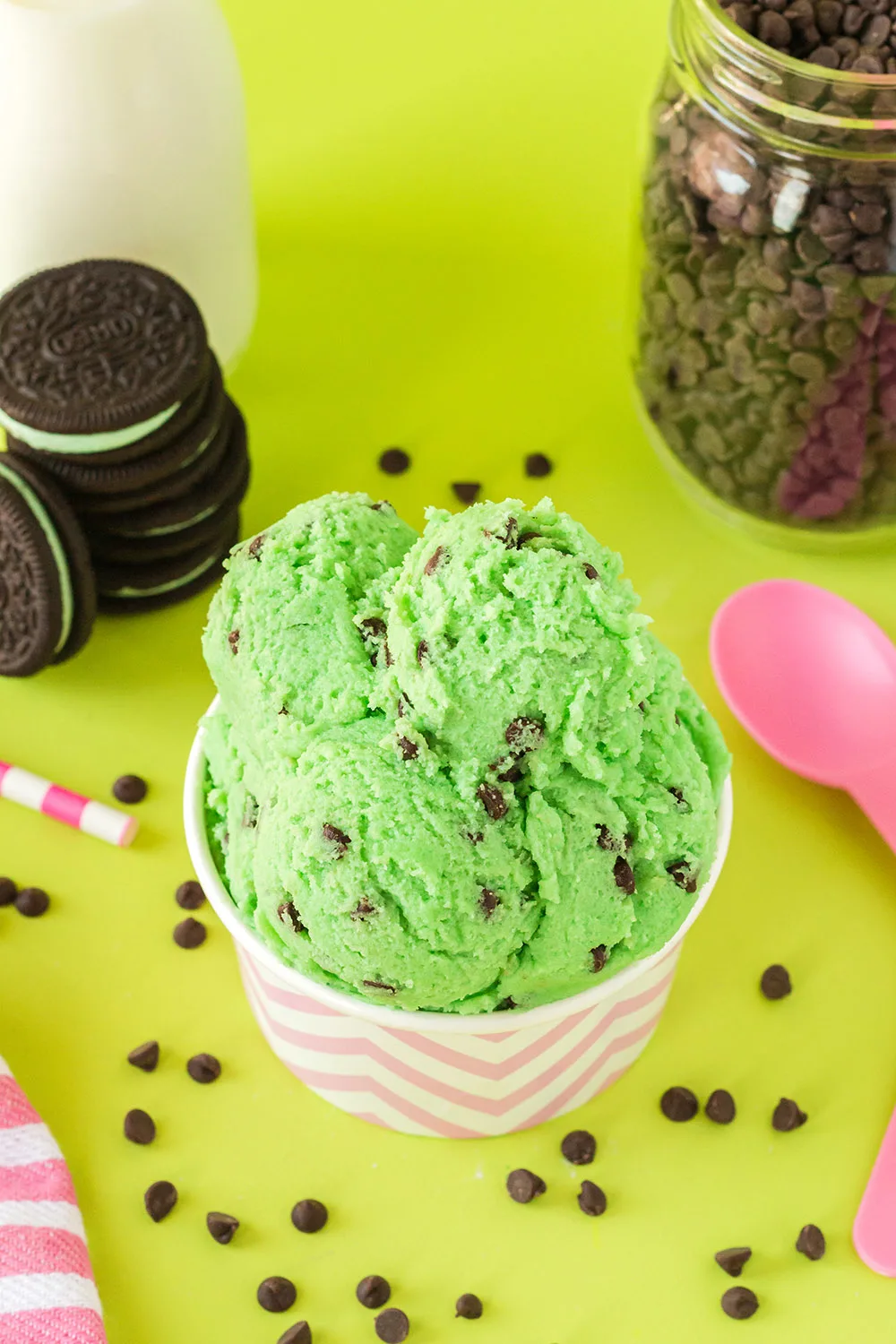 Scoops of mint chip dough in a pink cup with OREOs, chips, milk, and pink spoons on a green background.