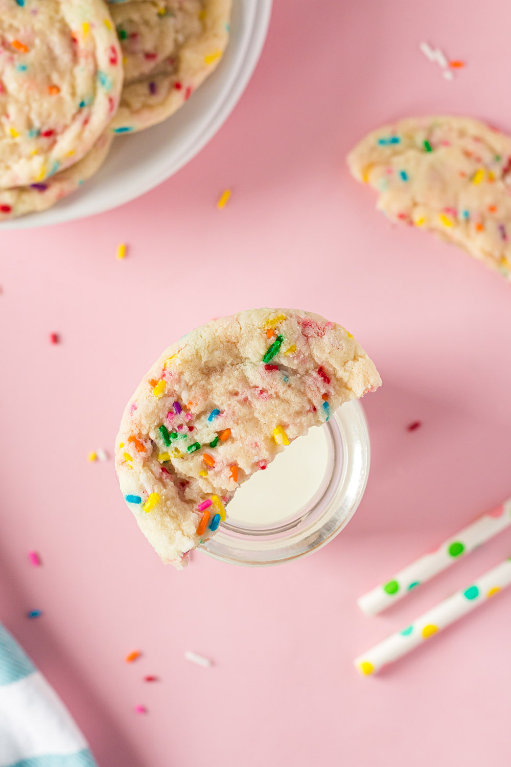Half of a confetti cookie on top of a glass of milk next to straws and more cookies.