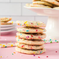 Stack of sugar cookies with rainbow sprinkles next to cookies on a stand and plates.
