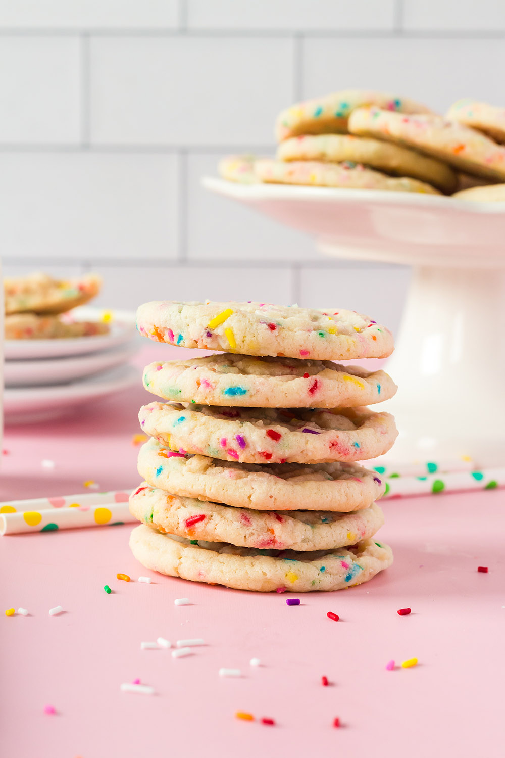 Stack of sugar cookies with rainbow sprinkles next to cookies on a stand and plates. 