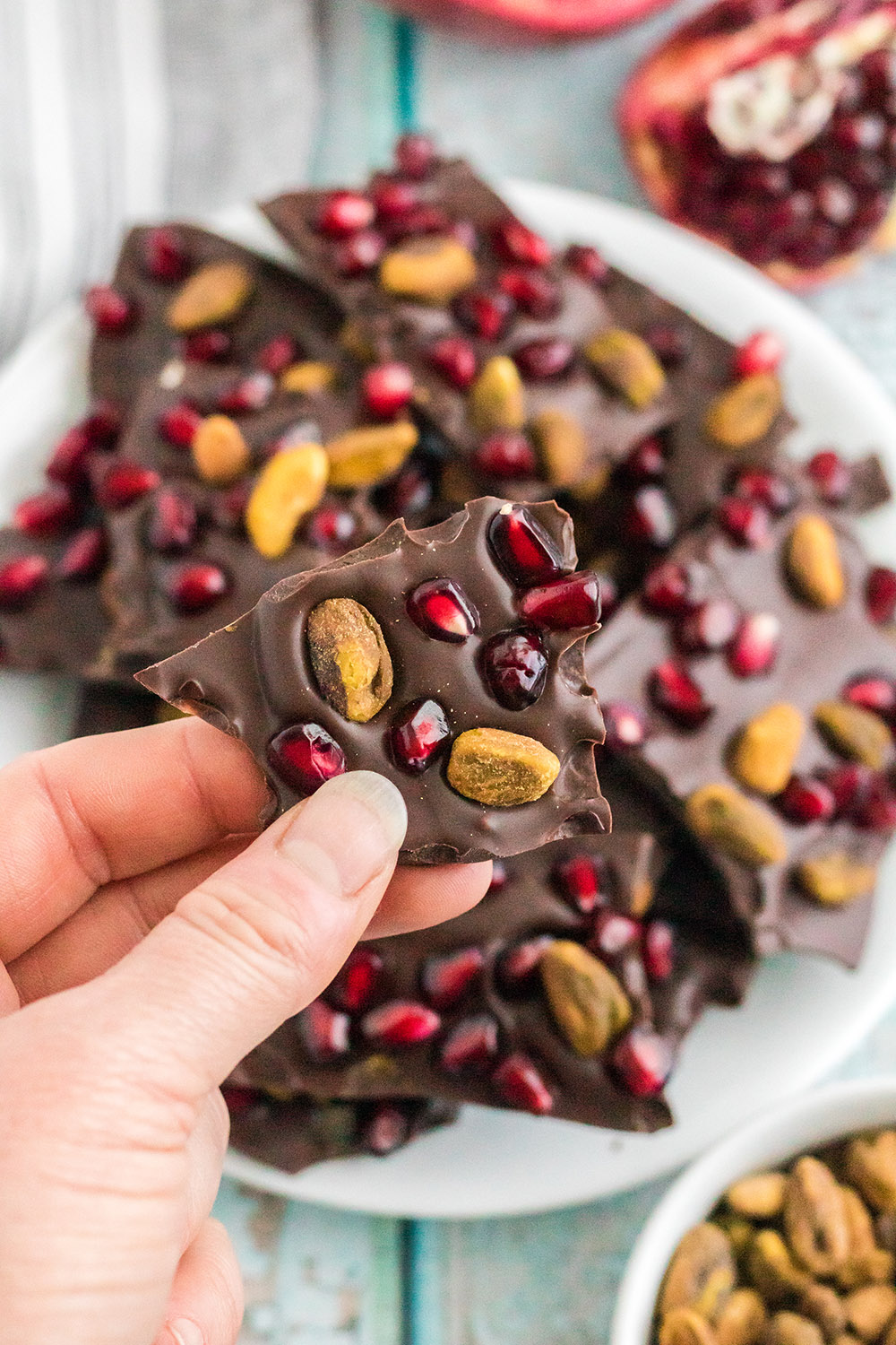 Holding a piece of chocolate pomegranate with pistachios candy over the plate full of the pieces. 