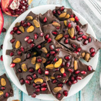 Chocolate bark candy with pomegranates and pistachios on a plate next to nuts and pomegranates on the table.