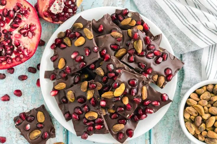 Chocolate bark candy with pomegranates and pistachios on a plate next to nuts and pomegranates on the table.