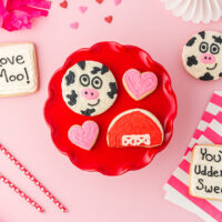 Valentine sugar cookies on a red plate. One cow, one barn, two hearts, with other cookies on the table.