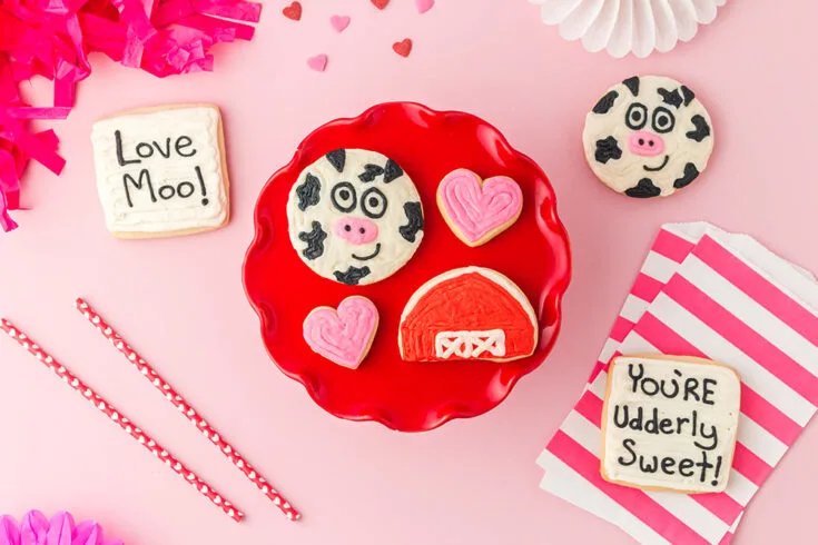 Valentine sugar cookies on a red plate. One cow, one barn, two hearts, with other cookies on the table.