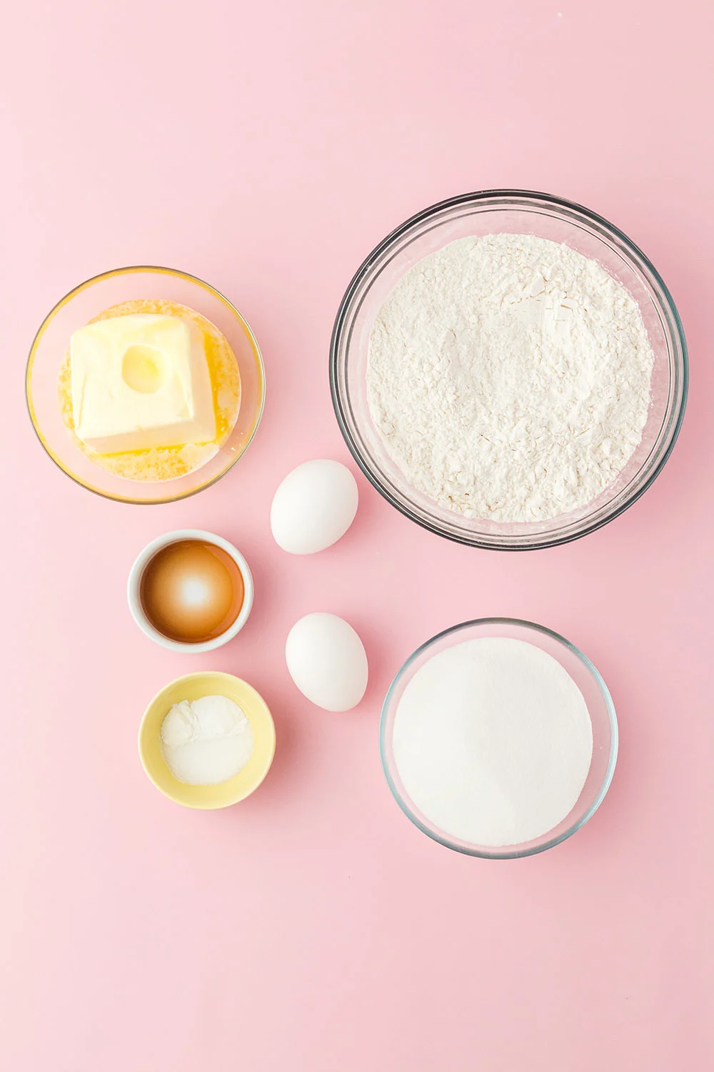 Butter, eggs, vanilla, flour, and baking powder in bowls on a pink table. 