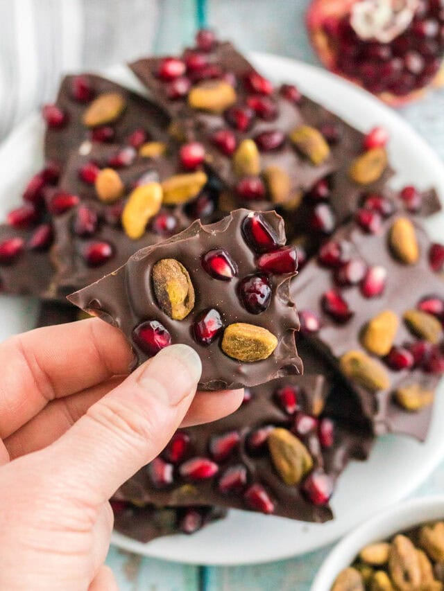 Holding a piece of chocolate pomegranate with pistachios candy over the plate full of the pieces.