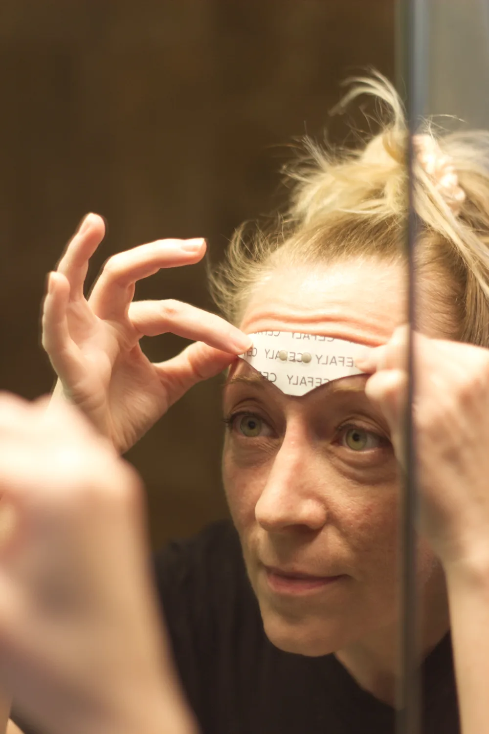 Girl placing a Cefaly electrode pad on her forehead in the mirror.