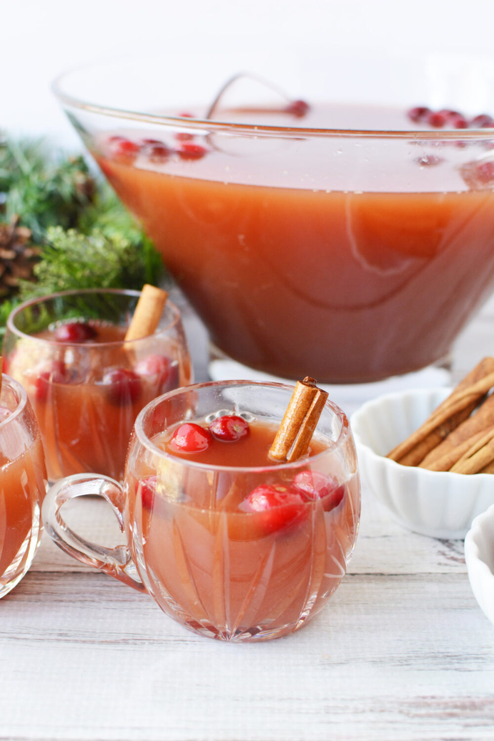 Mugs of punch with cranberries and cinnamon sticks next to the bowl of punch. 