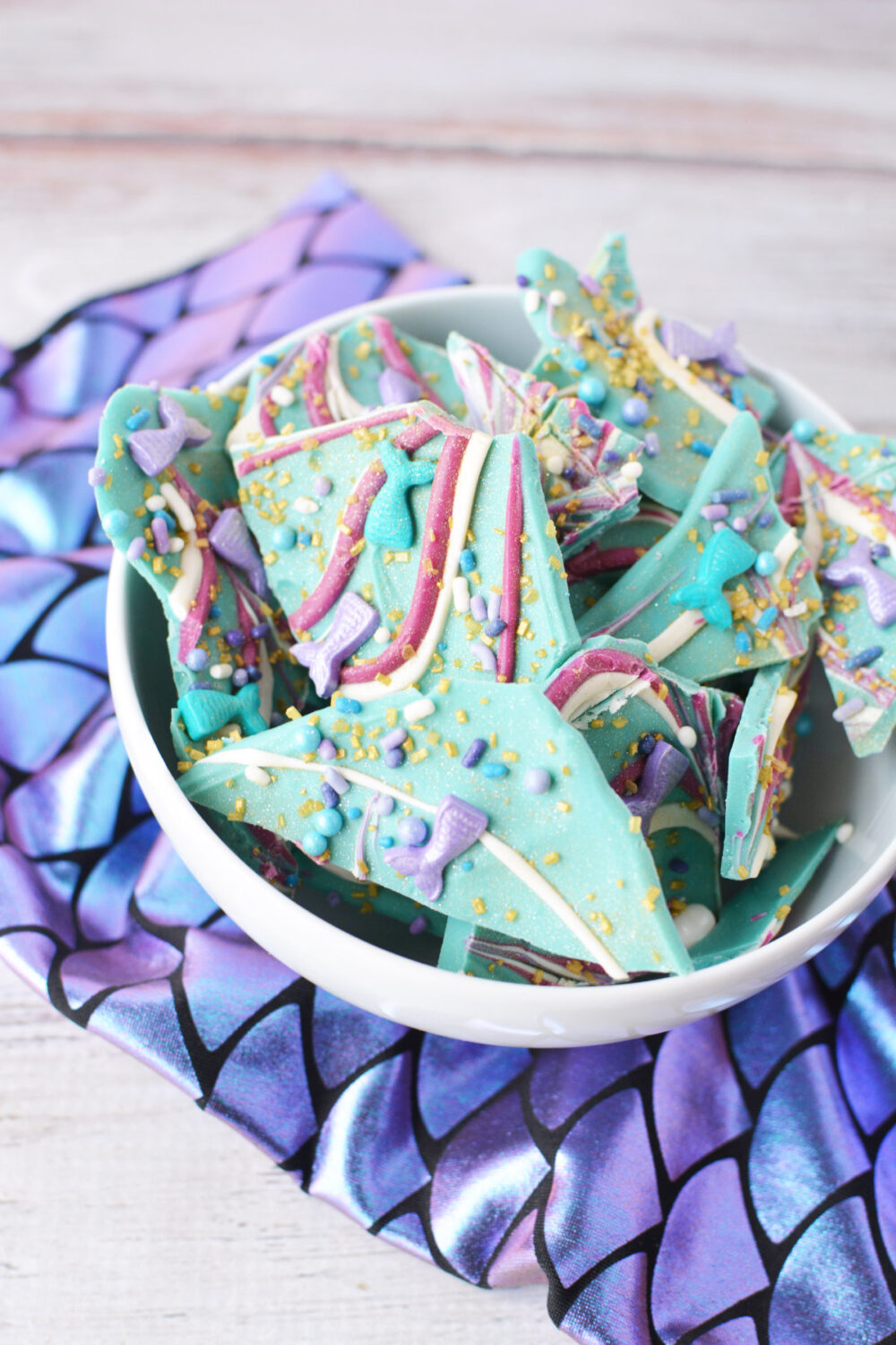 Bowl of mermaid candy bark pieces on top of mermaid fin scale fabric. 