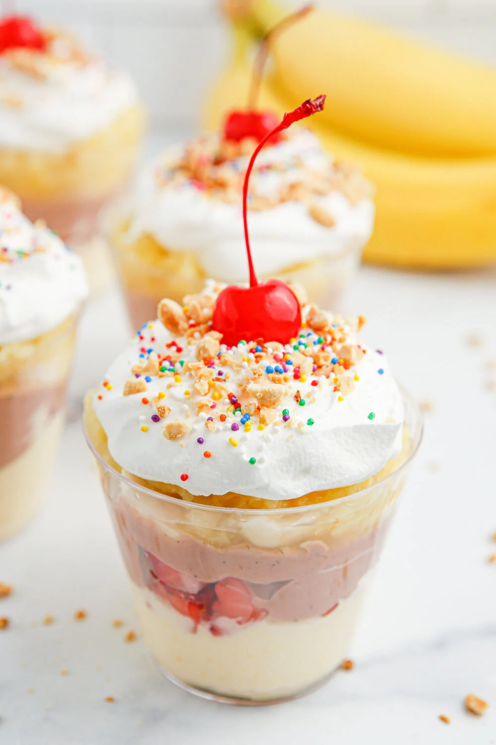 Banana split pudding parfait cups topped with whipped cream, cherries, and crushed peanuts. 