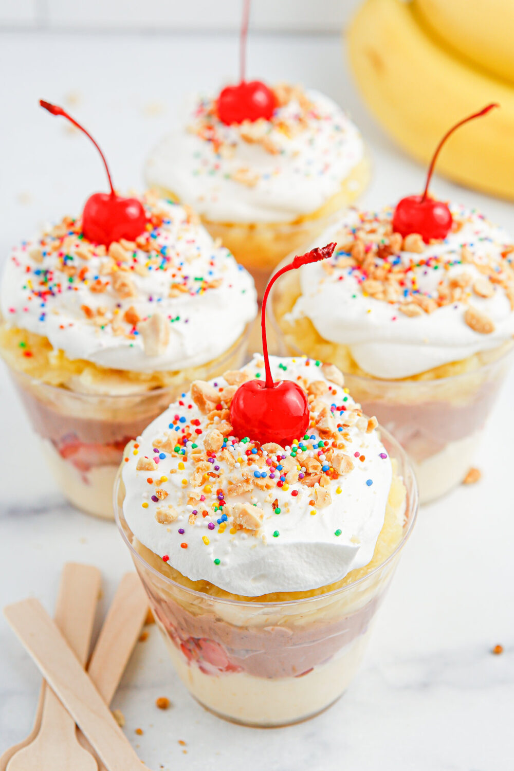 Four banana split pudding cups with whipped cream, sprinkles, and cherries. 