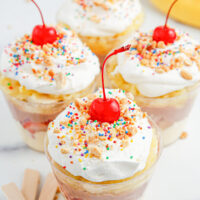Four banana split pudding cups with whipped cream, sprinkles, and cherries.