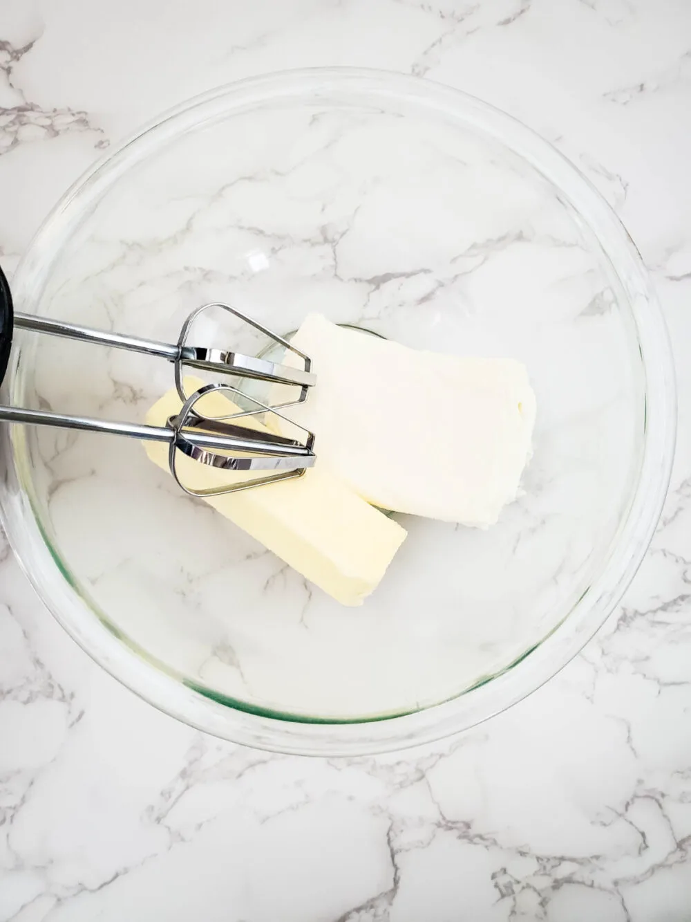 Butter and cream cheese in a clear mixing bowl with a mixer. 