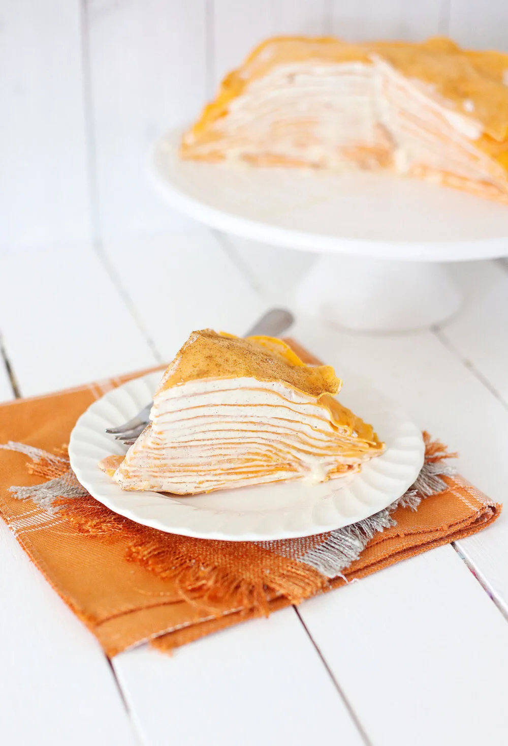 Slice of pumpkin crepe cake on a plate with the rest of the cake on a platter in the background. 