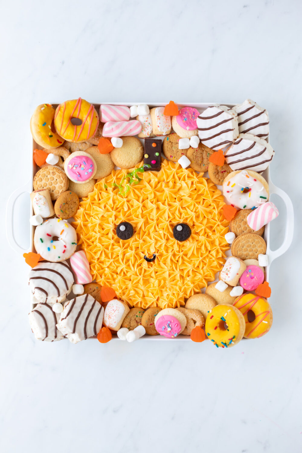 Cakes, candies, donuts, and cookies around an icing pumpkin on a board. 