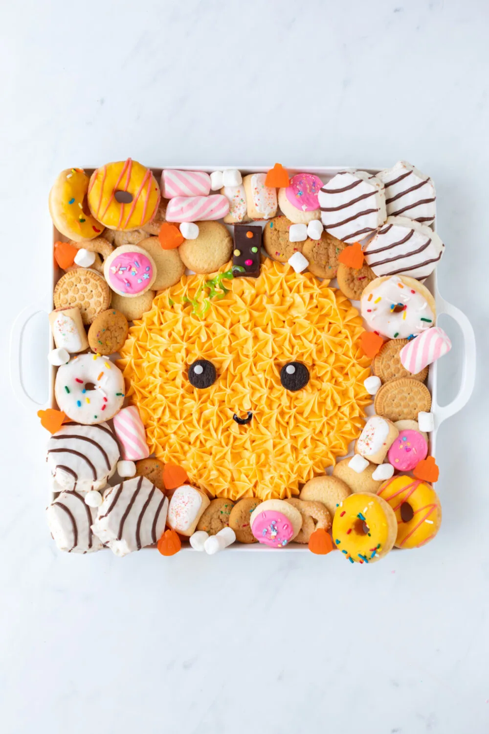 Cakes, candies, donuts, and cookies around an icing pumpkin on a board. 
