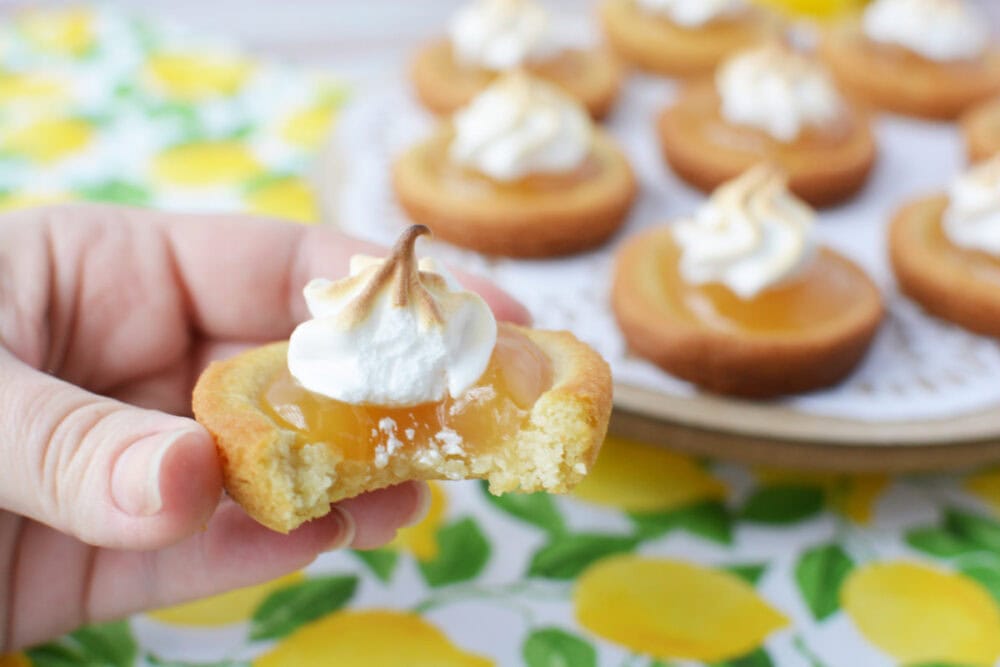 Holding a lemon meringue pie cookie with a plate full of them in the background.