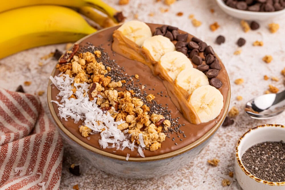 Bowl filled with chocolate peanut butter smoothie topped with banana slices, granola, coconut shreds, and more. 