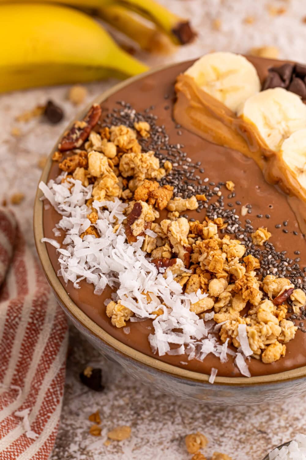 Chocolate smoothie bowl topped with coconut, granola, chia seeds, peanut butter, banana slices, and chocolate chips. 