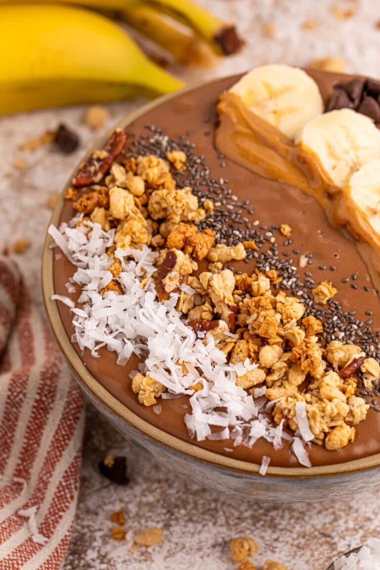 Chocolate smoothie bowl topped with coconut, granola, chia seeds, peanut butter, banana slices, and chocolate chips.