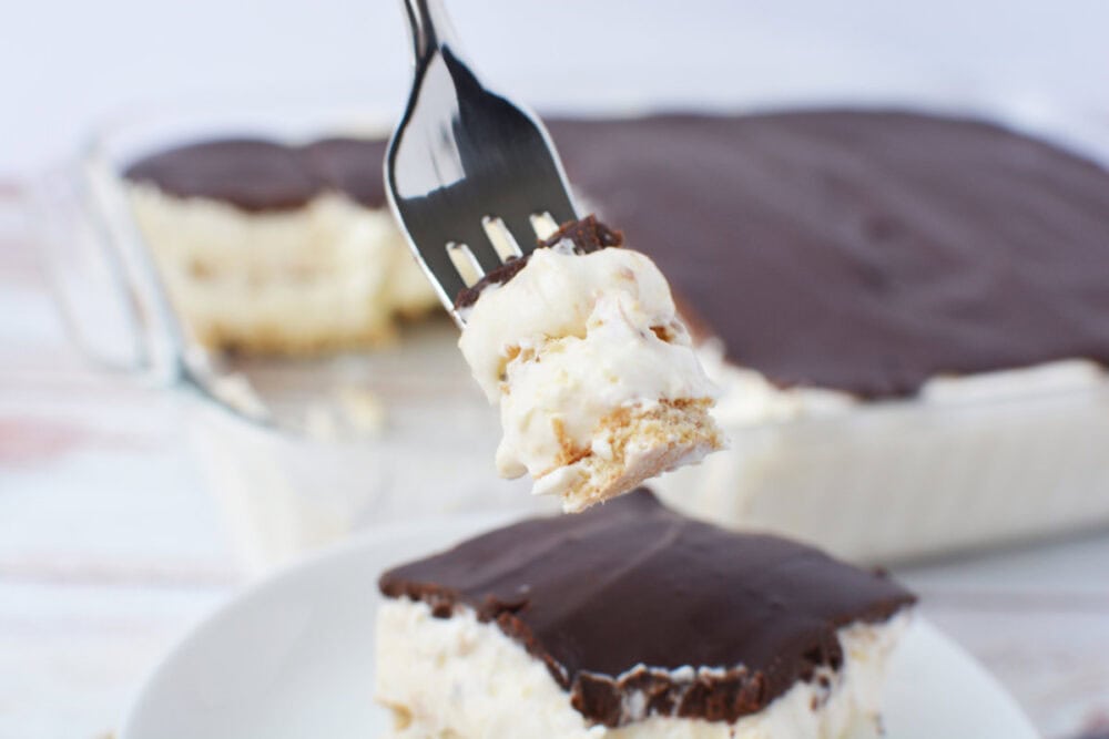 Fork with a bite of chocolate eclair cake on it above the slice and the whole cake in the background.