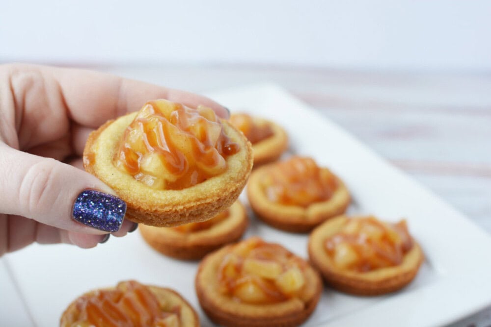 Holding a caramel apple pie cookie with the plate of them in the background. 