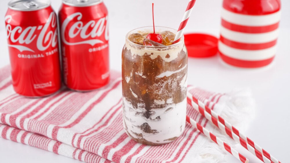 Glass of Fluffy Coke next to cans of Coca-Cola Classic and straws. 