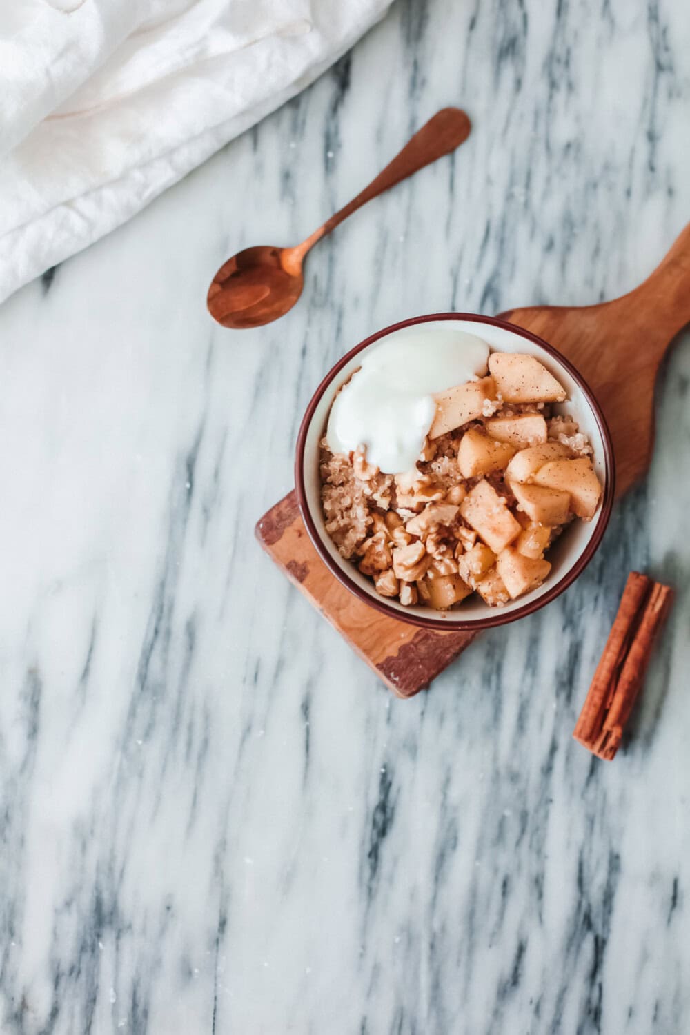 Breakfast bowl of apple and quinoa on a board next to a spoon and cinnamon stick. 