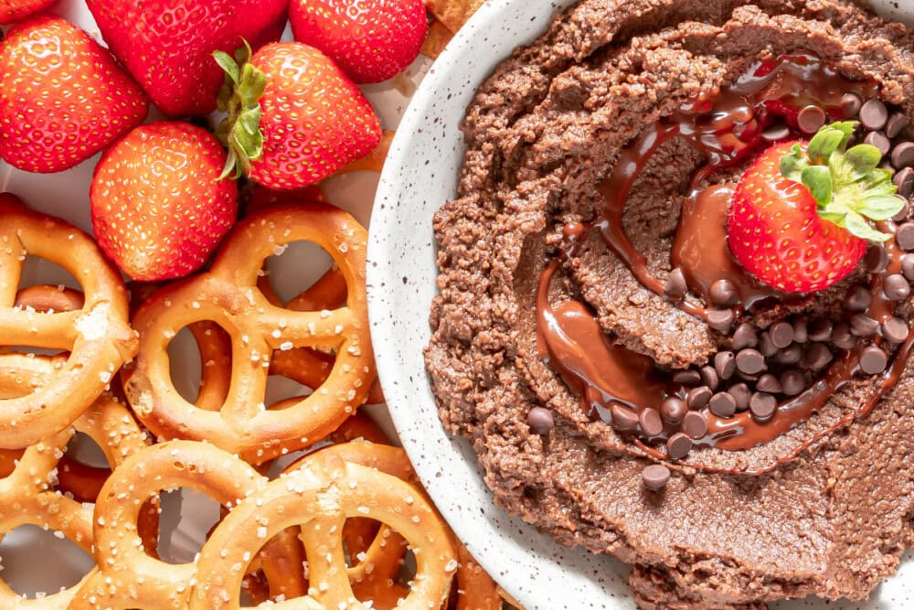 A bowl of chocolate hummus with a strawberry in it next to pretzels and strawberries. 