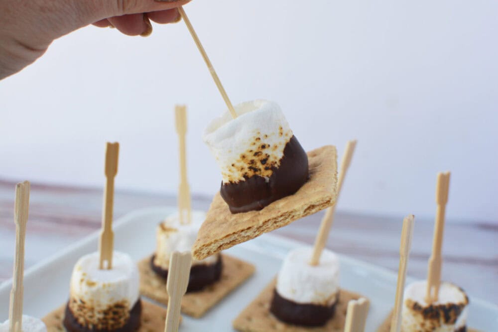 Holding a s'mores skewer with the rest on a tray in the back. 