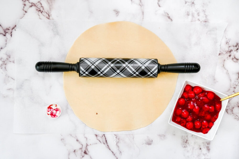 A rolling pin on pie crust next to cherry pie filling. 